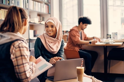 Two young female students have a conversation over a laptop and textbook in a university library cafe