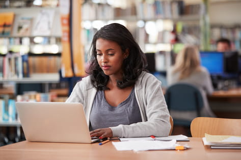 A female student works on her laptop at a table in the university library with her study notebook