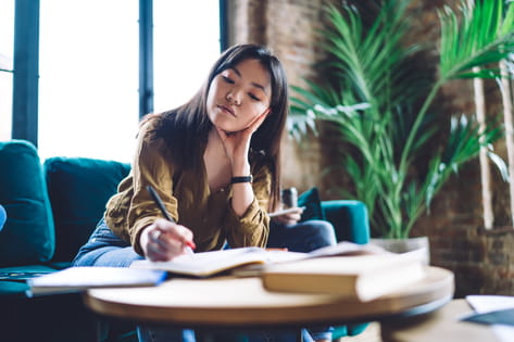 A female student sits on a blue sofa and writes into an open notebook on a small table