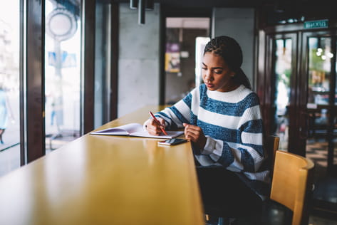 A female student sits at a high table in a university cafe writing in a notebook and looking at a mobile phone