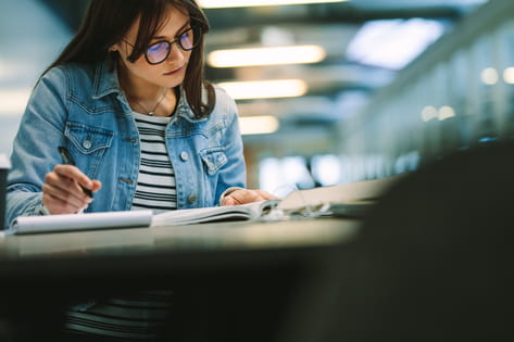 A young female student wearing glasses reads a textbook and writes in a notebook on the table in a university library