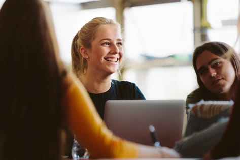 A group of three female students smile as they work on a project together over a laptop