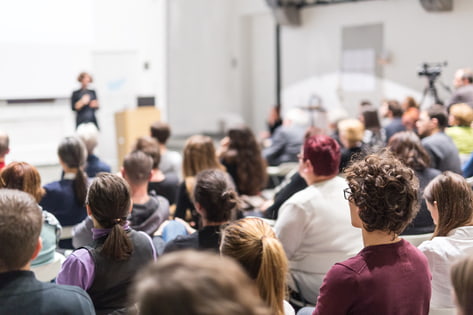 A professor teaches a large university class in a lecture theatre as the audience listens intently