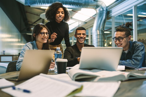 Four students surround a single laptop in a university library and smile with open books on the table