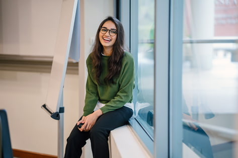 A smiling female student sits on a windowsill in a seminar room with a presentation board behind her