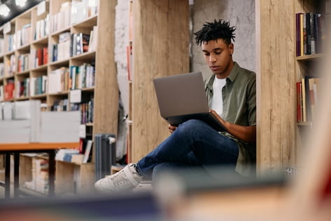 A young male student works on his laptop amongst bookshelves in a university library