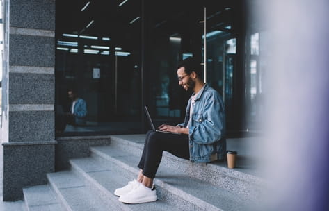 A male student sits outside on the university library steps while typing on his laptop