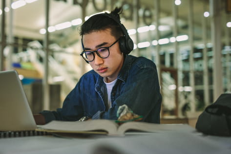 A male student wears headphones while watching his laptop in the university library in the evening