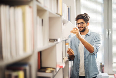 A male student smiles as he takes a book from a university library bookshelf